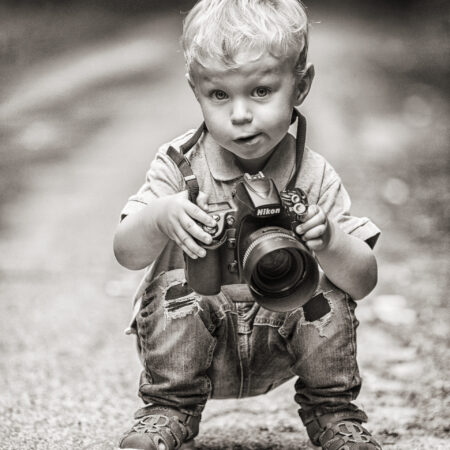 Ein kleiner Junge mit blonden Locken hält eine große Kamera in den Händen und schaut neugierig in die Kamera. Das Bild ist in Schwarz-Weiß gehalten und fängt einen besonderen Moment bei einem Kinder-Fotoshooting im Fotostudio Sabrina Zeuge in Unna ein.